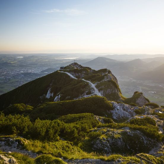 Aerial view of a mountain peak with a winding road leading to the top, surrounded by lush greenery and rocky terrain. The horizon shows distant mountains and a city under a clear sky.