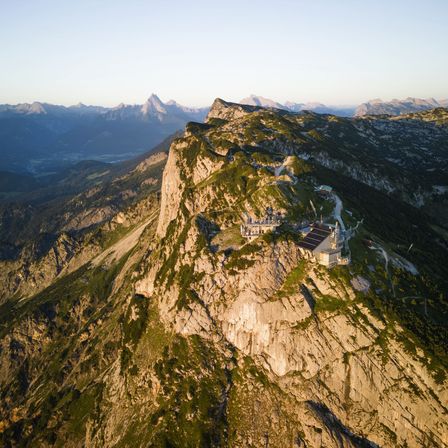 Ein atemberaubender Luftblick auf einen Klippenrand mit einem Gebäude oben, umgeben von einem üppigen grünen Tal und majestätischen Bergen.