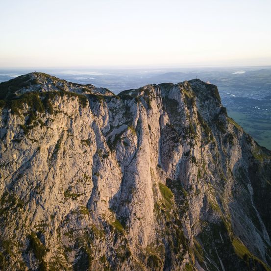 Aerial view of a rocky cliff face with rugged terrain, sparse vegetation, and a distant landscape under a clear sky.