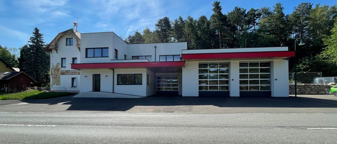 Ein weißes Gebäude mit rotem Rand befindet sich in einer ländlichen Umgebung. Das Gebäude verfügt über große Fenster und eine Garage mit Glastüren. Es ist von Bäumen und einem klaren blauen Himmel umgeben.