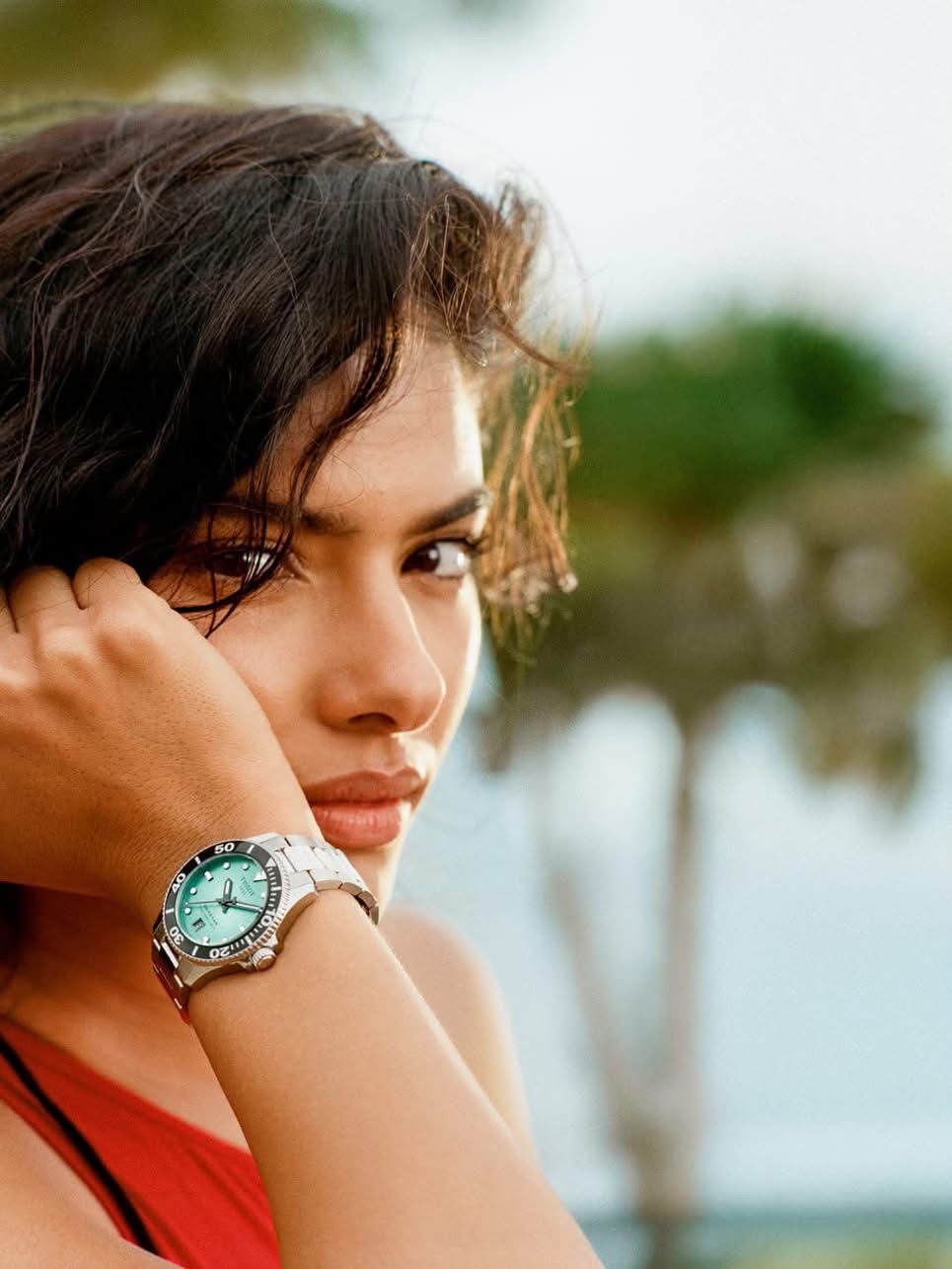 Eine Frau mit dunklem Haar schaut auf ihre Uhr, die einen türkisen Ziffernblatt und ein silbernes Armband hat.