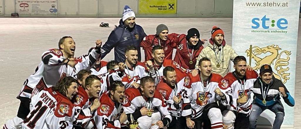 Eine Gruppe von Eishockeyspielern feiert mit ihrem Pokal auf einer Eisbahn, mit Zuschauern im Hintergrund.
