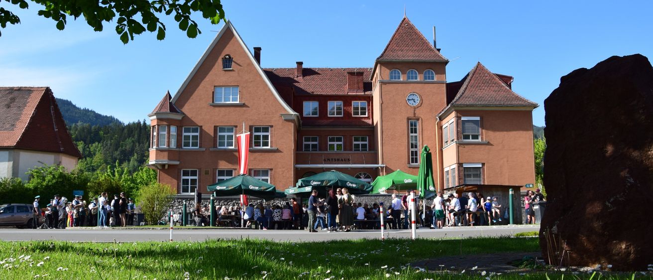 Ein großes Gebäude mit einem Uhrturm, umgeben von grünen Sonnenschirmen, beherbergt eine Menschenmenge draußen an einem sonnigen Tag.