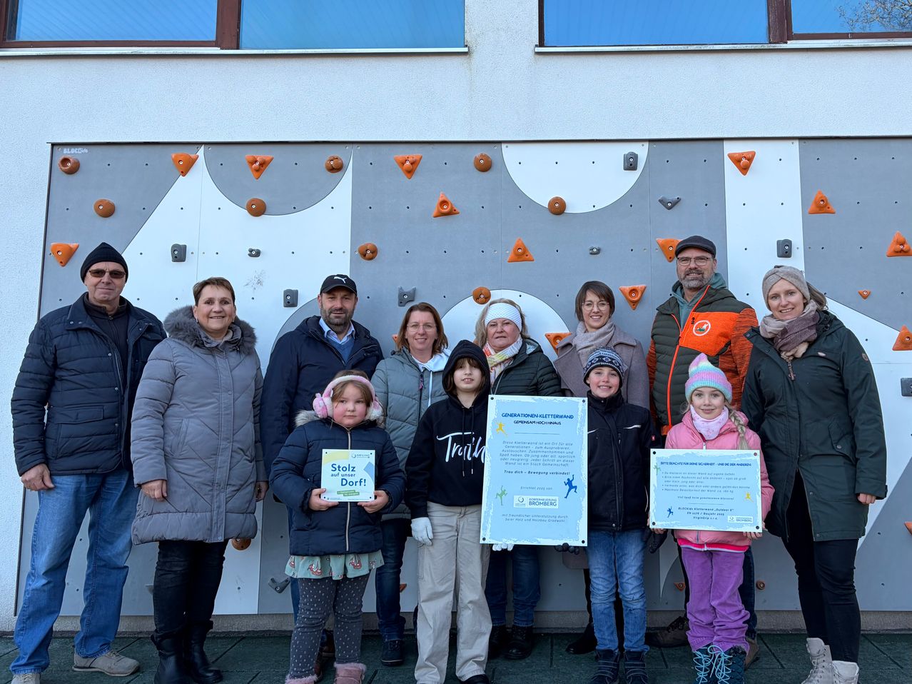 Eine Gruppe von Menschen, darunter Kinder, steht gemeinsam vor einer Kletterwand und hält Urkunden in den Händen. Sie lächeln für ein Foto.