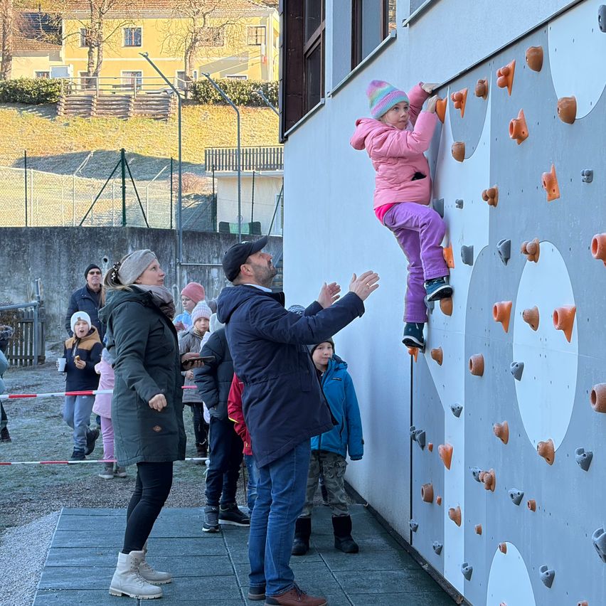 Ein kleines Mädchen in einer rosa Jacke klettert an einer Außenwand. Ein erwachsener Mann in einer schwarzen Kappe feuert sie an. Mehrere Menschen, darunter Kinder, stehen herum und beobachten.