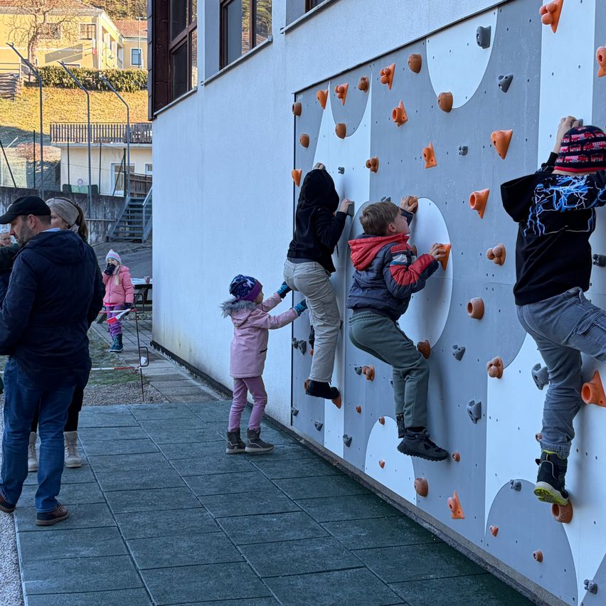 Kinder klettern an einer Wand mit verschiedenen Griffen, während andere zusehen. Ein Mann und eine Frau stehen in der Nähe, einer trägt einen Hut und die andere eine Mütze.