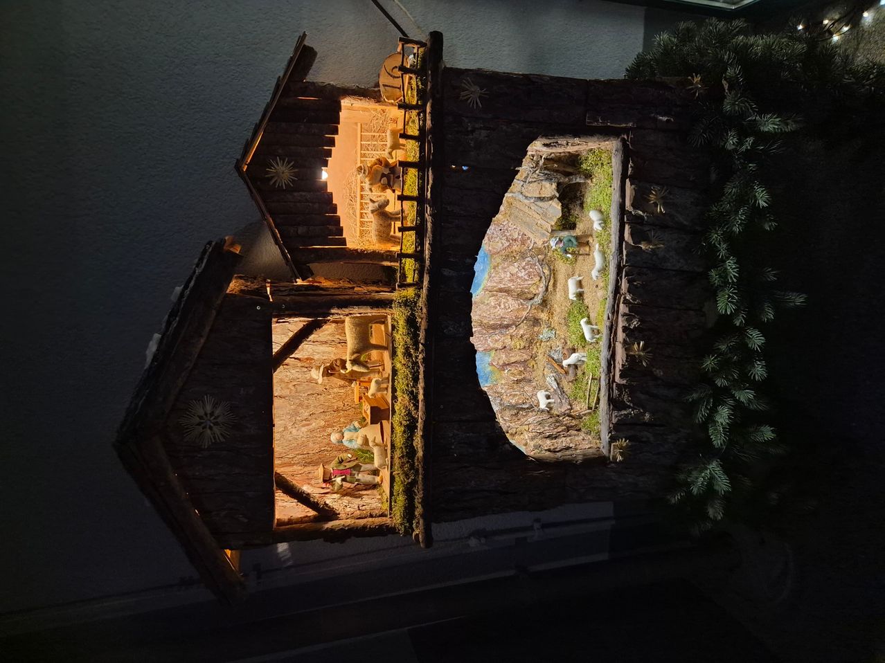 Auf einem dunklen Hintergrund zeigt eine hölzerne Krippe eine beleuchtete Scheune. Figuren von Jesus, Maria und Josef sind sichtbar. Draußen gibt es einen felsigen Hügel mit einem Grasbereich und Schafen.