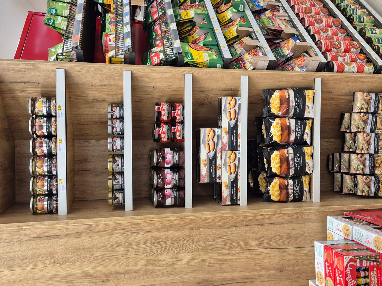 Ein Holzregal zeigt verschiedene Lebensmittelprodukte, einschließlich Dosenwaren, Kartons und Tüten mit Nudeln, in organisierten Abschnitten angeordnet.