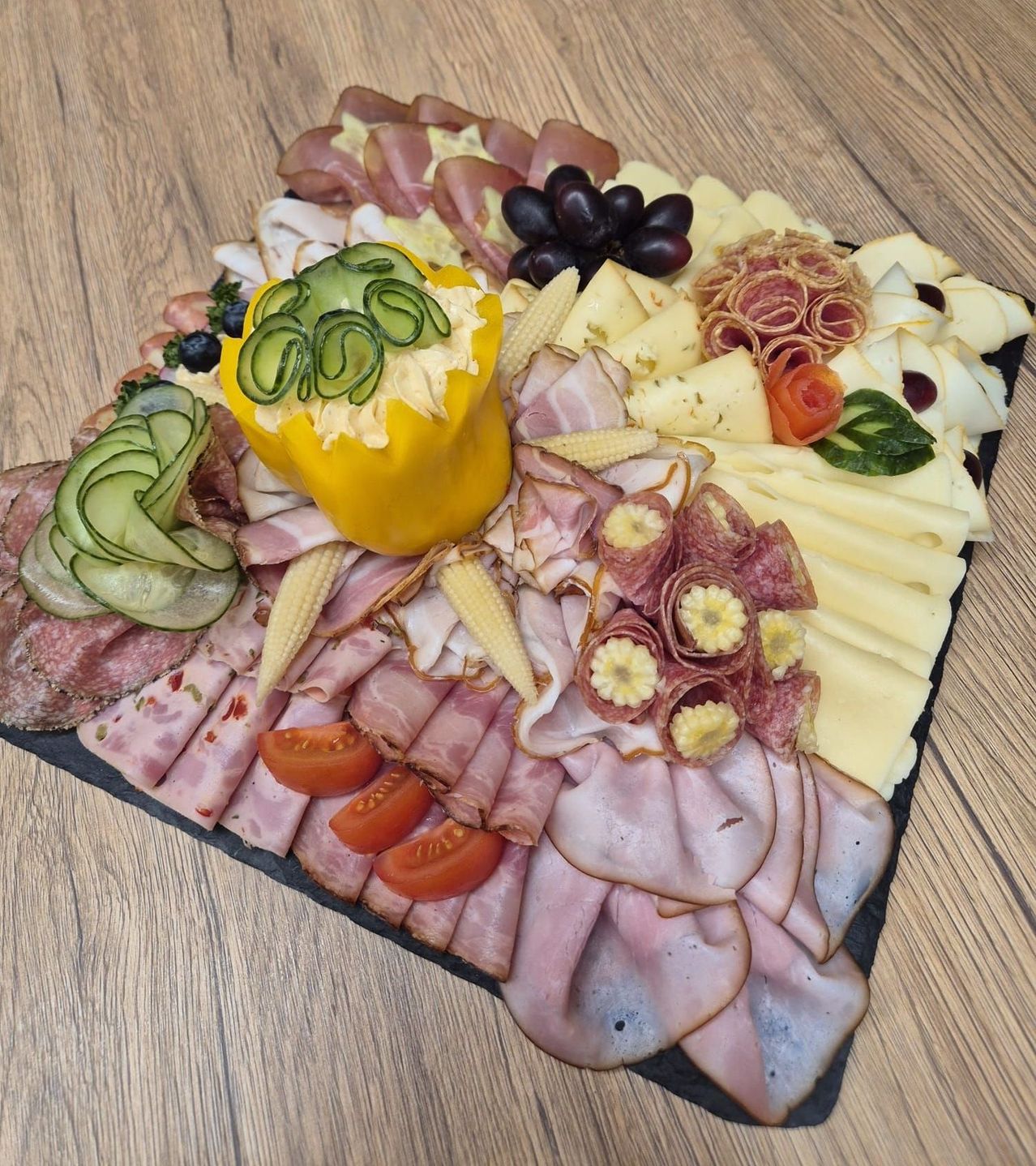 Ein Charcuterie-Brett mit einer Auswahl an Fleisch, Käse und Gemüse. Es enthält geschnittenen Schinken, Salami und Wurstwaren, begleitet von Tomaten, Gurken und Mais.