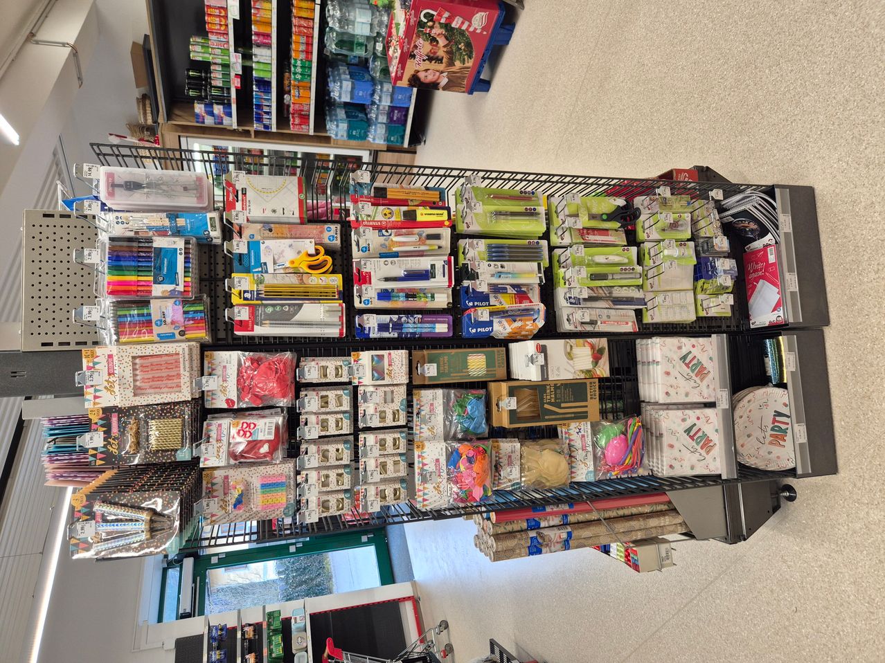 Ein Ladengang mit einer Vielzahl bunter Produkte zur Schau. Artikel umfassen Stifte, Bleistifte, Scheren und andere Schreibwaren. Es gibt auch Geschenkboxen, Geschenkpapier und Bänder.