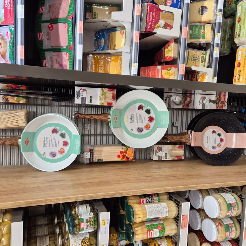 Ein hölzernes Ladenregal mit verschiedenen Lebensmitteln, darunter Bratpfannen und Gläser. Die Pfannen haben Etiketten mit Lebensmittelillustrationen. Es gibt auch verpacktes Fleisch und Käse.