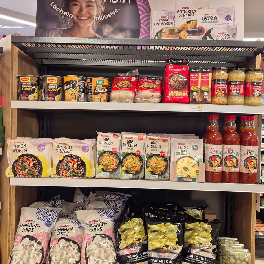 Ein Regal in einem Laden zeigt verschiedene asiatische Lebensmittel. Es gibt Ramen-Nudeln, Nudelbecher, Saucen und Chips. Das obere Regal hat Dosenartikel, und das untere Regal hat verpackte Artikel. Ein Poster im Hintergrund zeigt eine lächelnde Frau.
