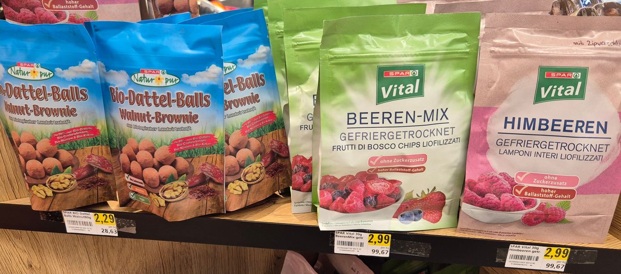 Ein Regal zeigt zwei Beutel mit getrockneten Fruchtsnacks. Ein Beutel ist mit 'el-Balls Brownie' beschriftet und zeigt ein farbenfrohes Bild von Nüssen und Beeren. Der andere Beutel ist mit 'Vital' beschriftet und zeigt eine Mischung aus gefrorenen Beeren mit einem grünen Etikett.