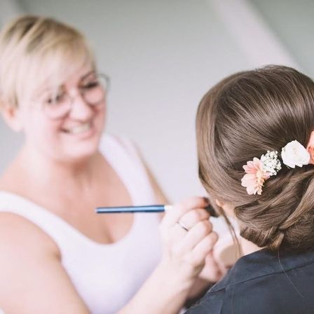 Eine Frau mit Brille lächelt, während sie Make-up auf das Haar einer anderen Frau aufträgt, die eine Haarsträhne mit Blumen-Haarschmuck hat.