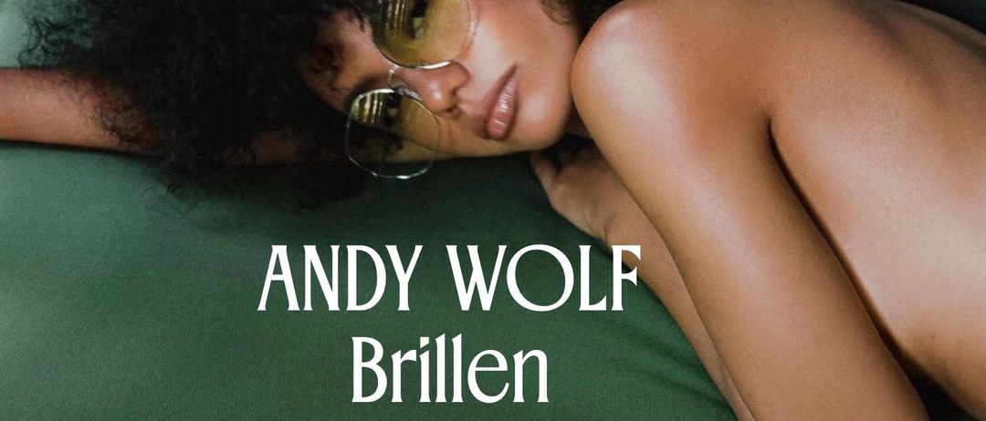 Eine Frau mit lockigem Haar und Sonnenbrille liegt auf einer grünen Couch mit dem Kopf auf der Armlehne. Das Bild hat einen Textüberlagerung, der 'Andy Wolf Brillen POP-UP Freitag, 31. Juli' lautet.