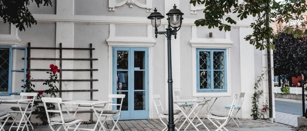 Ein weißes Gebäude mit blauen Fenstern und einer blauen Tür. Draußen sind Tische und Stühle aufgestellt, in der Nähe sind Blumen.