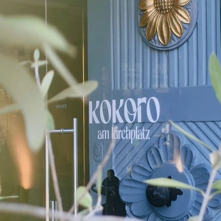 Eine blaue Tür mit dem Wort Kokoro in weißen Buchstaben über einem goldenen Sonnenblumendesign. Die Tür ist mit einem schwarzen Griff und einem Schloss verziert.