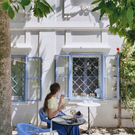 Eine Frau sitzt an einem Tisch vor einem weißen Gebäude mit blauen Fenstern und hält ein Glas Wein. Die Umgebung ist hell und sonnig, mit dekorativen Lichtern und viel Grün.
