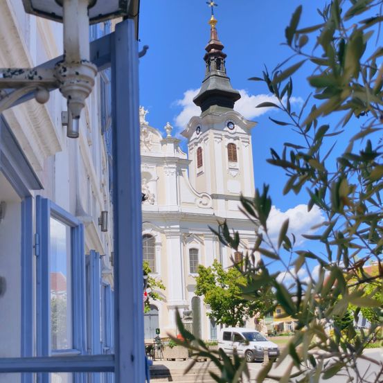 Eine weiße Kirche mit einem hohen Turm und Uhren ist durch einen blauen Fensterrahmen zu sehen. Bäume und ein geparkter weißer Van sind im Vordergrund.