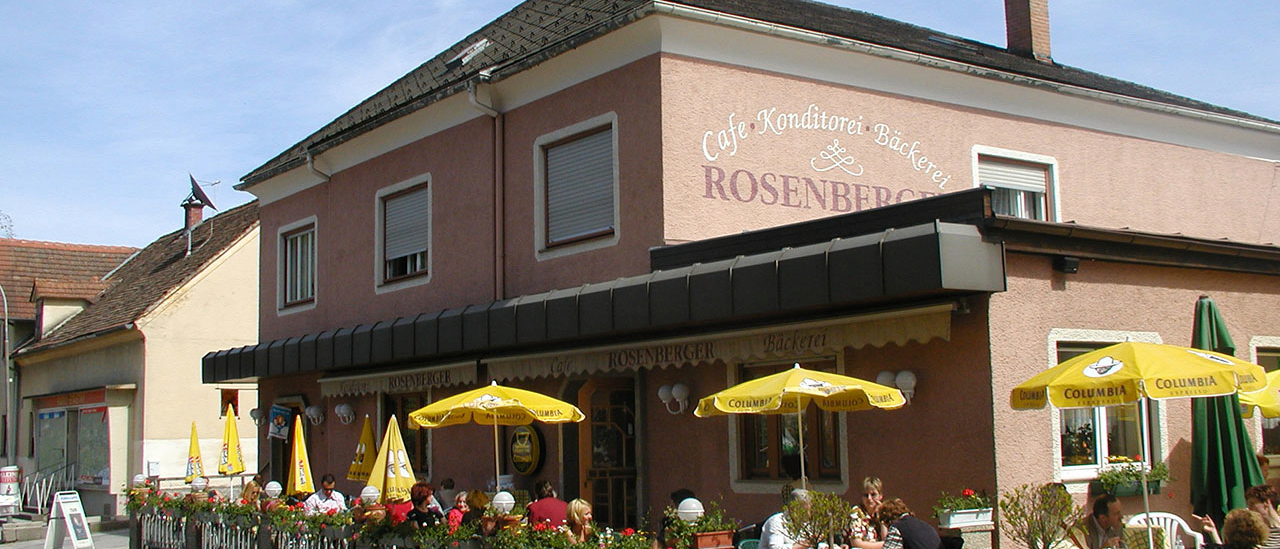 Ein Café mit einer rosa Fassade und dem Namen Cafe Konditorei Backerei Rosenberger. Es sitzen Menschen draußen unter gelben Sonnenschirmen.
