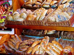 Ein hölzerne Regal zeigt mehrere Korbkörbe, die mit verschiedenen Arten von Brot gefüllt sind, darunter Brezeln, Sesam-Röllchen und andere. Das Brot ist ordentlich angeordnet, und die Körbe sind übereinander gestapelt.