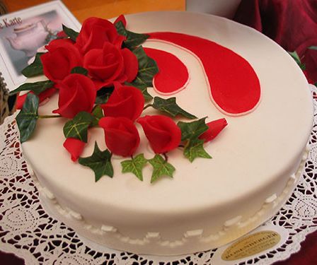 Ein weißer Kuchen mit roten Rosen und grünen Blättern auf einer Spitzendeckchen. Der Kuchen hat ein rotes Wirbeldesign.