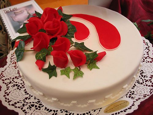 Ein weißer Kuchen mit roten Rosen und grünen Blättern auf einer Spitzendeckchen. Der Kuchen hat ein rotes Wirbeldesign.