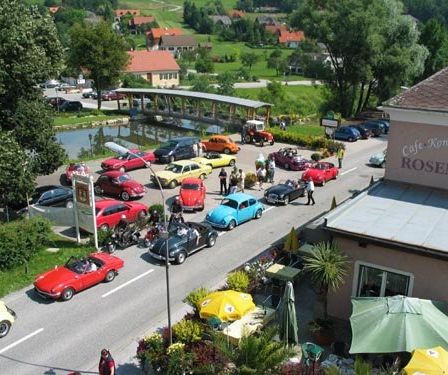 Eine Vogelperspektive eines Parkplatzes mit Oldtimern, die am Straßenrand geparkt sind, in der Nähe eines Cafés mit dem Namen 'Café Konditorei Rosenbauer'. Es gibt Leute, die herumlaufen, und eine Brücke in der Ferne.