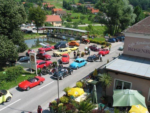 Eine Vogelperspektive eines Parkplatzes mit Oldtimern, die am Straßenrand geparkt sind, in der Nähe eines Cafés mit dem Namen 'Café Konditorei Rosenbauer'. Es gibt Leute, die herumlaufen, und eine Brücke in der Ferne.