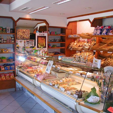 Eine Bäckerei mit einer Glasvitrine voller verschiedener Gebäcke und Brote. An den Seiten befinden sich Regale mit Snacks und Getränken. Die Decke hat Lichter und Lüftungsöffnungen.