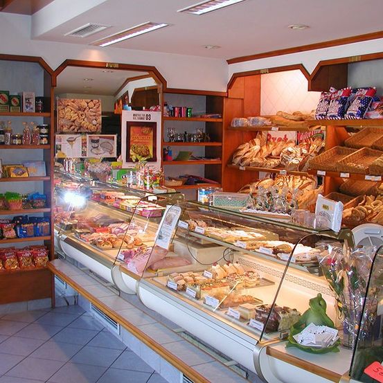 Eine Bäckerei mit einer Glasvitrine voller verschiedener Gebäcke und Brote. An den Seiten befinden sich Regale mit Snacks und Getränken. Die Decke hat Lichter und Lüftungsöffnungen.