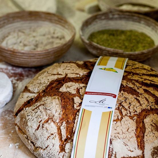 Ein Laib rustikales Brot mit einem Etikett darauf, auf einer Holzoberfläche platziert, mit Mehl und anderen Zutaten im Hintergrund.