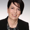 Eine Frau mit kurzen, dunklen Haaren und einem schwarzen Blazer lächelt in die Kamera und zeigt ihre Zähne. Sie hat helle Haut und trägt ein gemustertes Oberteil.