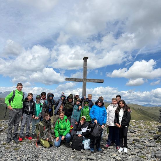 Eine Gruppe von Menschen in Jacken und Mützen, die lächelnd für ein Foto posieren, ist auf einem Berg mit einem Kreuz im Hintergrund.