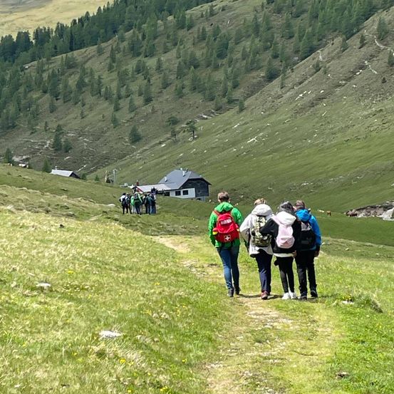 Eine Gruppe von Wanderern geht einen grasbewachsenen Bergpfad hinauf, mit Bäumen und einem Haus in der Ferne. Ein Wanderer trägt einen roten Rucksack.