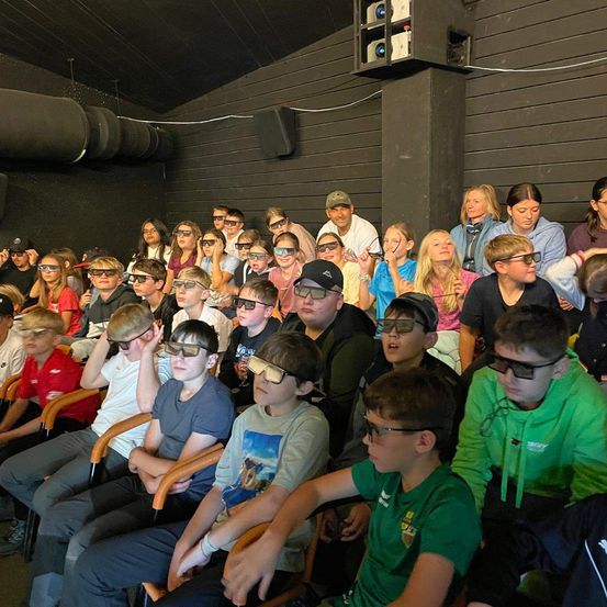 Eine große Gruppe von Menschen, die 3D-Brillen tragen, sitzen in einem schwach beleuchteten Raum, einige stehen, wahrscheinlich bei der Betrachtung eines 3D-Films oder einer Präsentation.