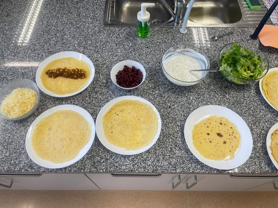 Eine Küchenarbeitsplatte mit verschiedenen Lebensmitteln: mehrere Teller mit gelben Tortillas, eine Schüssel mit roten Bohnen, eine Schüssel mit Dressing und eine Schüssel Salat. Im Hintergrund befindet sich ein Waschbecken mit Wasserhahn und Seifenspender.