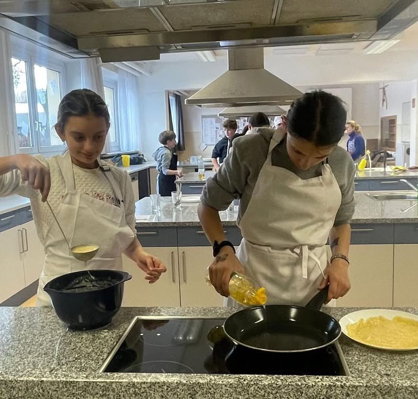 Zwei Personen in einer Küche, eine gießt Öl in eine Pfanne, während die andere einen Topf rührt. Verschiedene Küchenutensilien und Personen sind im Hintergrund zu sehen.