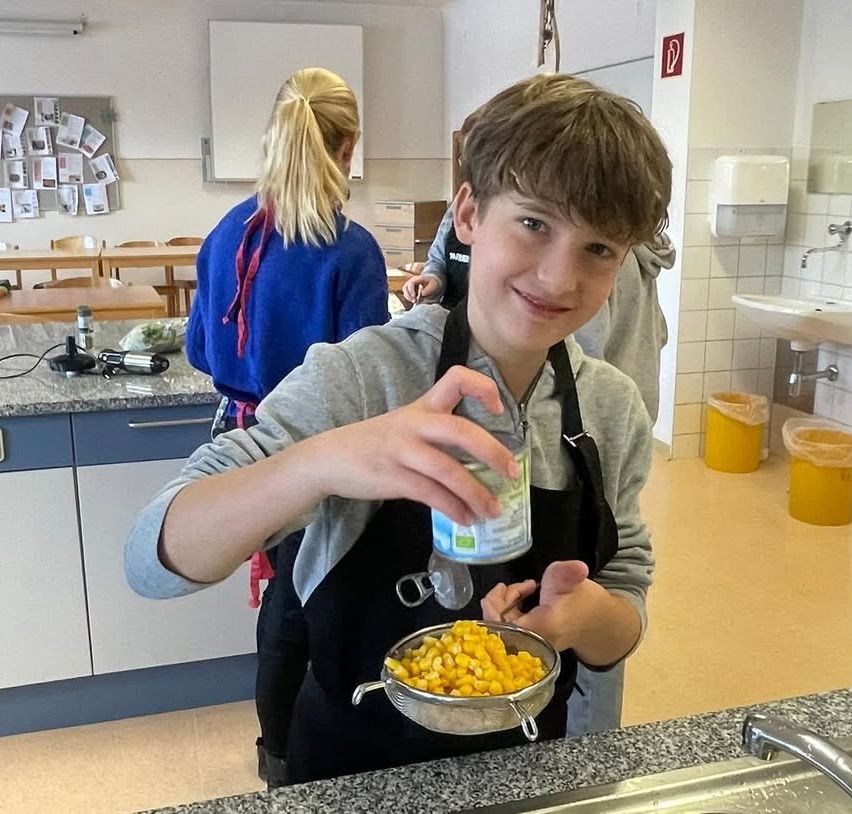 Ein Junge in einer Küche gießt Mais in einen Sieb, während eine Frau in einem blauen Shirt zuschaut. Der Raum hat eine weiße Wand mit einem Bulletin Board und einem Waschbecken.