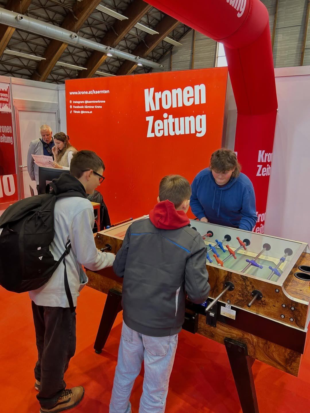 Mehrere Personen stehen um einen Tischfußballtisch in einem Gebäude. Der Banner hinter ihnen lautet 'Kronen Zeitung'.