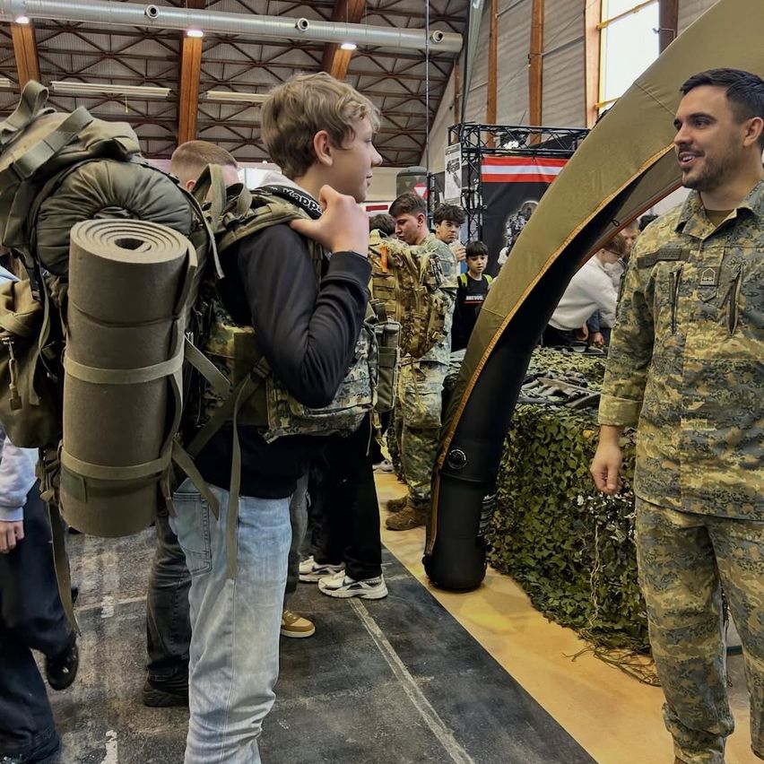 Ein junger Mann mit Rucksack unterhält sich mit einem Militäroffizier. Sie befinden sich in einem Raum mit militärischer Einrichtung, einschließlich getarnter Zelte und militärischer Ausrüstung.