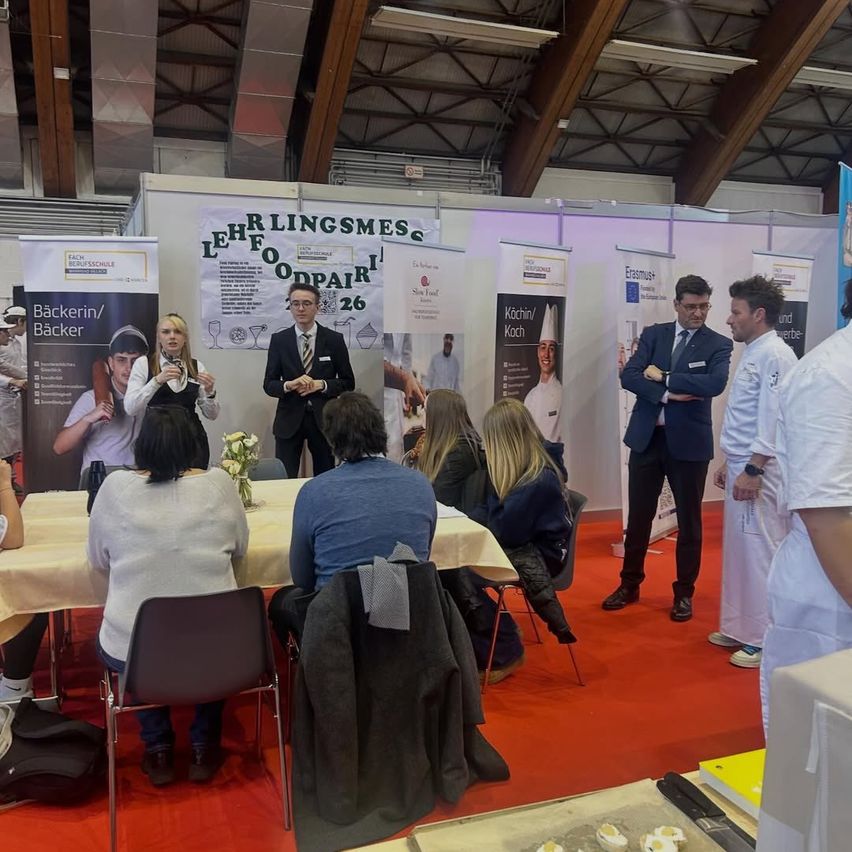Eine Gruppe von Menschen sitzt um einen Tisch herum, während ein Mann vor ihnen steht. Der Mann hält eine Präsentation und das Publikum hört aufmerksam zu. Dahinter wird ein Plakat mit einem Koch und den Worten "Lehrlingsmesse" angezeigt.