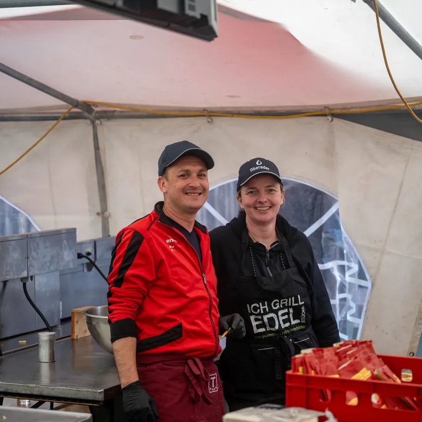 Zwei Personen lächeln in einer Küche, eine trägt eine rote Jacke und die andere ein schwarzes Apron. Ein roter Kasten mit Gegenständen steht in der Nähe.