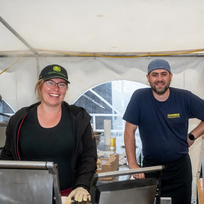 Ein Mann und eine Frau stehen in einem Zelt, lächeln und bereiten Essen zu. Der Mann trägt ein blaues T-Shirt und eine Uhr, und die Frau trägt eine schwarze John Deere-Kappe und eine Brille. Hinter ihnen befindet sich ein Fenster und ein Tisch mit Essen und einem Glas.