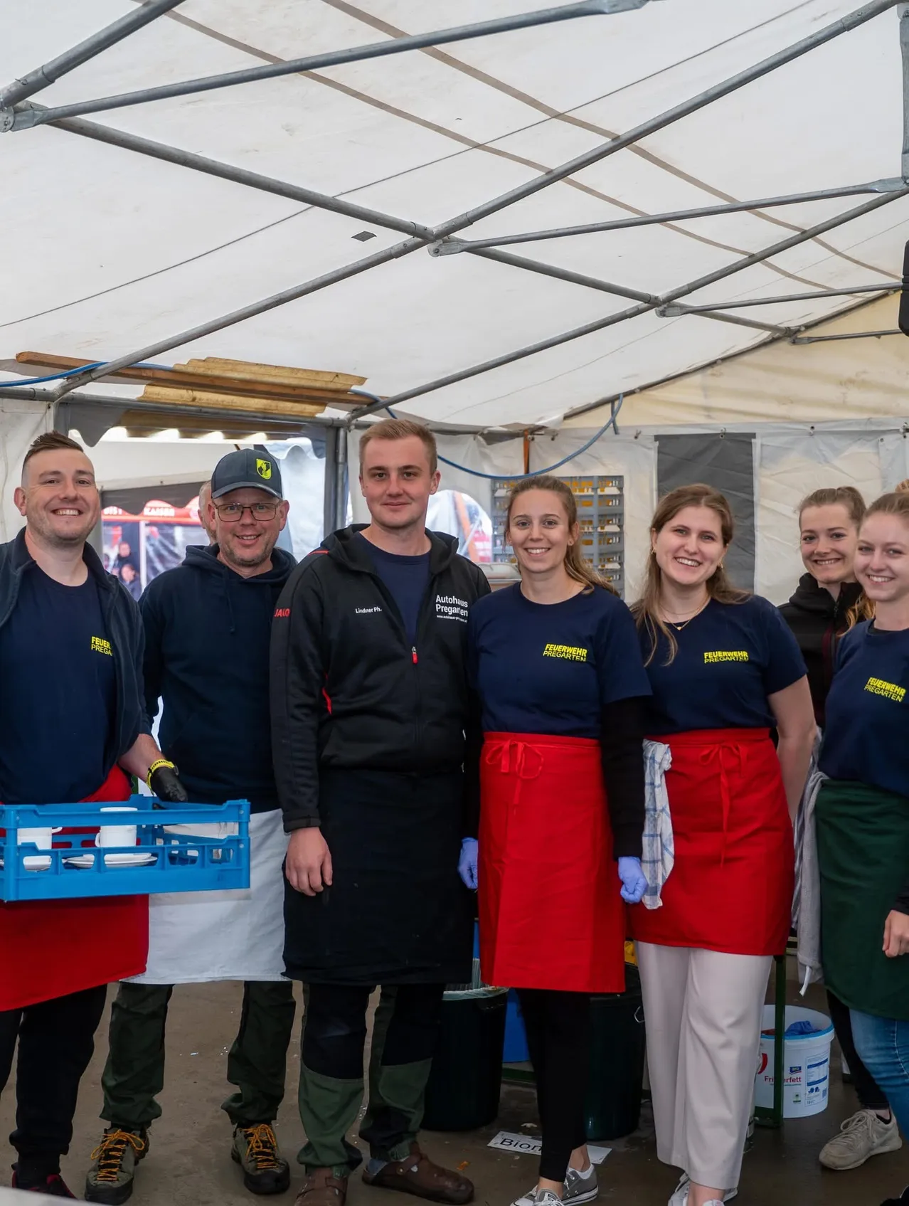 Eine Gruppe von Menschen in blauen Uniformen und Schürzen, die lächelnd und zusammenstehend unter einem Zelt stehen. Eine Person hält einen blauen Korb.
