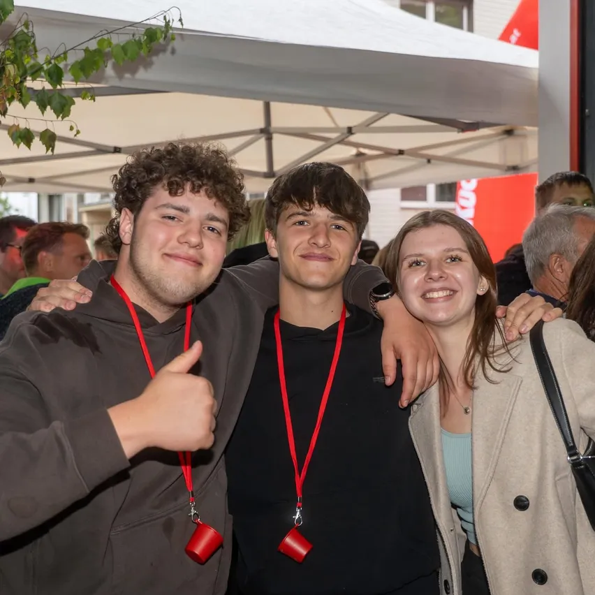 Drei junge Leute, zwei Männer und eine Frau, stehen eng beieinander unter einem Zelt und posieren für ein Foto. Sie tragen alle rote Lanyards.