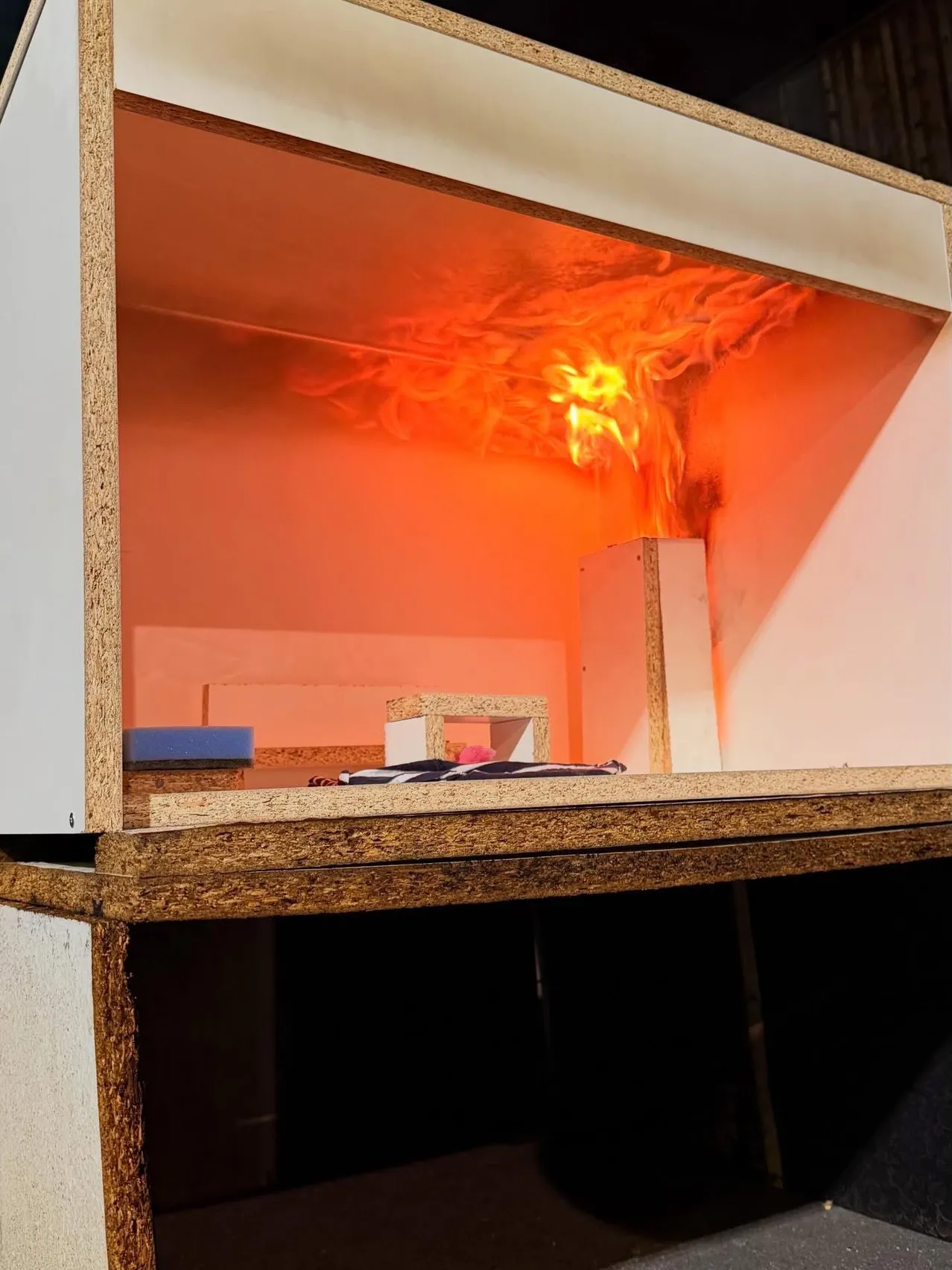 Ein Feuer brennt in einem kleinen Holzmodellkasten und erhellt den Raum mit einem orangefarbenen Schein. Der Kasten ist geöffnet und es gibt verschiedene Gegenstände verstreut.