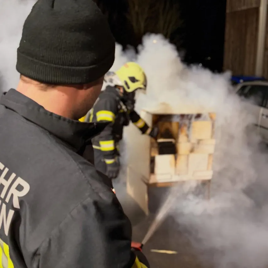 Ein Feuerwehrmann in Schutzkleidung sprüht Wasser auf einen brennenden Holzlagerkasten. Ein anderer Feuerwehrmann steht in der Nähe. Rauch füllt die Luft.