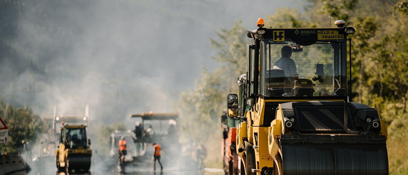 Ein Baustelle mit Arbeitern und Maschinen. Ein gelber und schwarzer BOMAG-Walze fährt mit Arbeitern in der Nähe die Straße entlang.