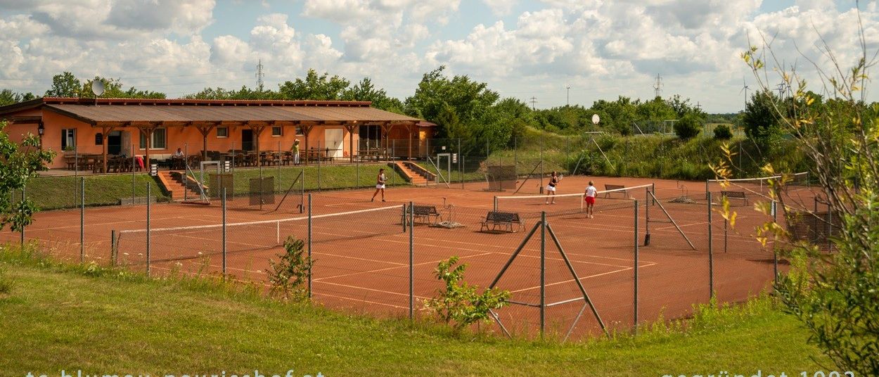 Tennisspieler auf einem Platz, mit einem Gebäude und Treppen im Hintergrund, an einem sonnigen Tag.